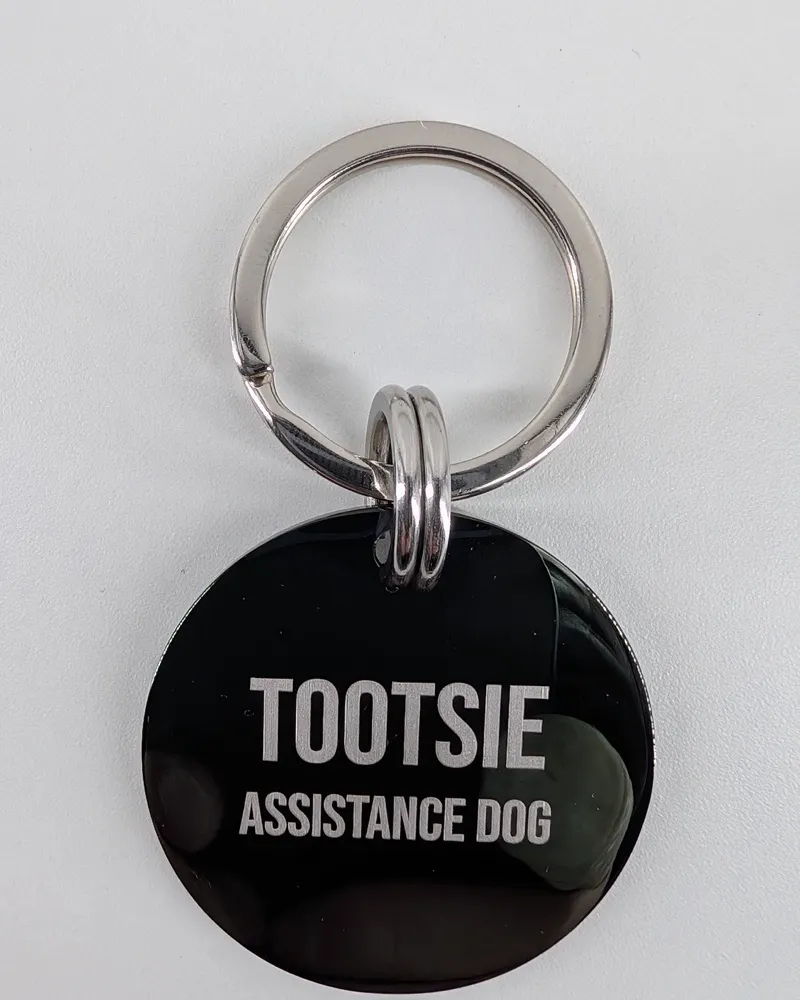 A black MoodTag round tag deep laser engraved TOOTSIE, ASSISTANCE DOG, on the Never-Fall Ring and the 26mm attachment ring